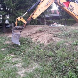 Artur-prace ziemne - Żółta koparka JCB w trakcie prac ziemnych w Olsztynie. Widoczny wykop i fragment domu w tle. Prace ziemne z bliska, zieleń i drzewa w otoczeniu.