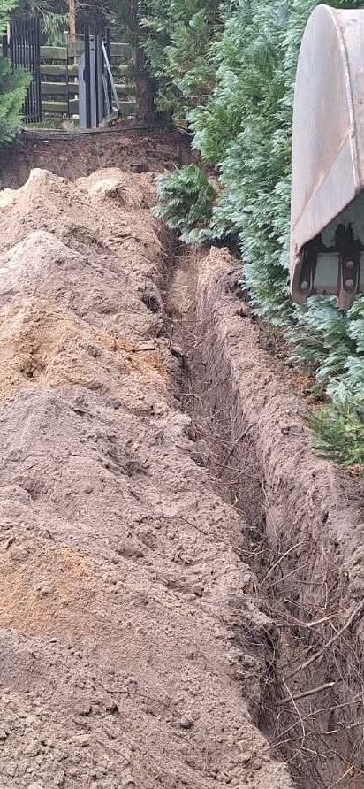 Wąski wykop w ziemi, widoczny fragment łyżki koparki, tło z zielonych krzewów i fragment ogrodzenia. Wykop świeży, z widocznymi korzeniami.