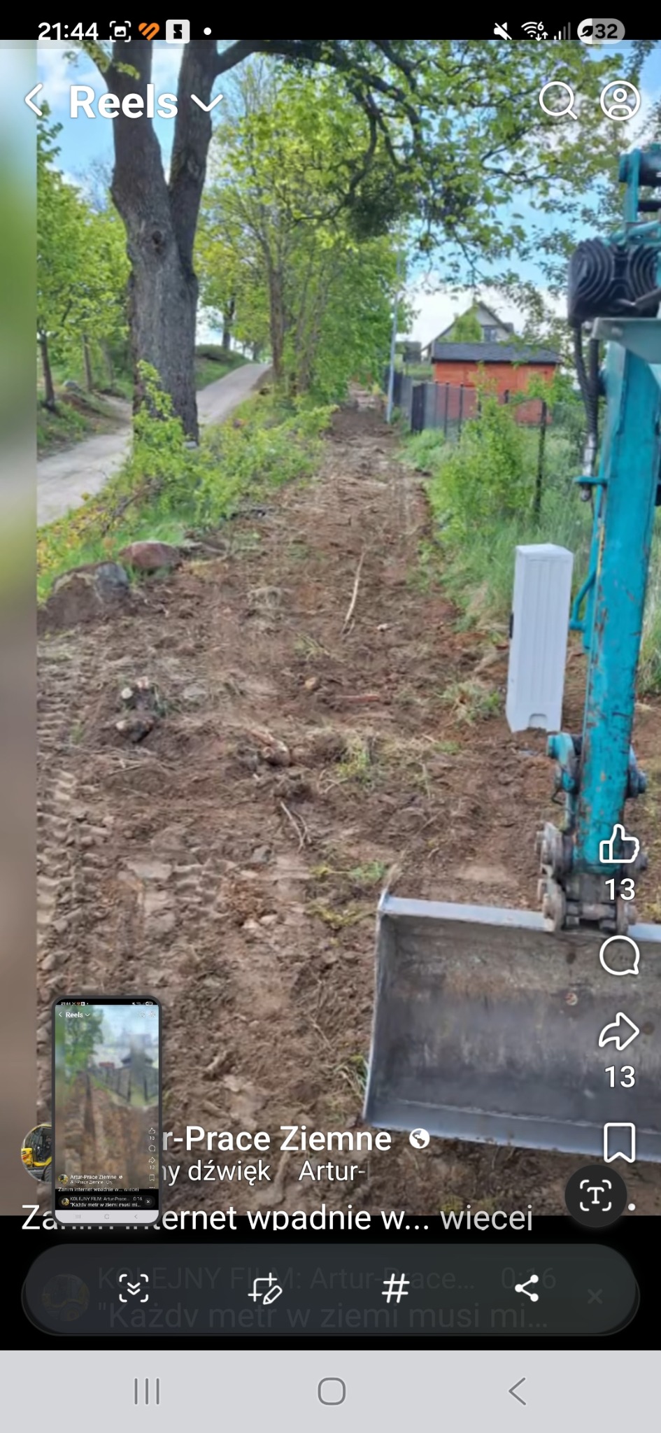 Wąska droga gruntowa po niwelacji, widoczna z perspektywy operatora koparki. Ślady opon i wycięte pnie drzew. W tle zabudowania i zieleń.