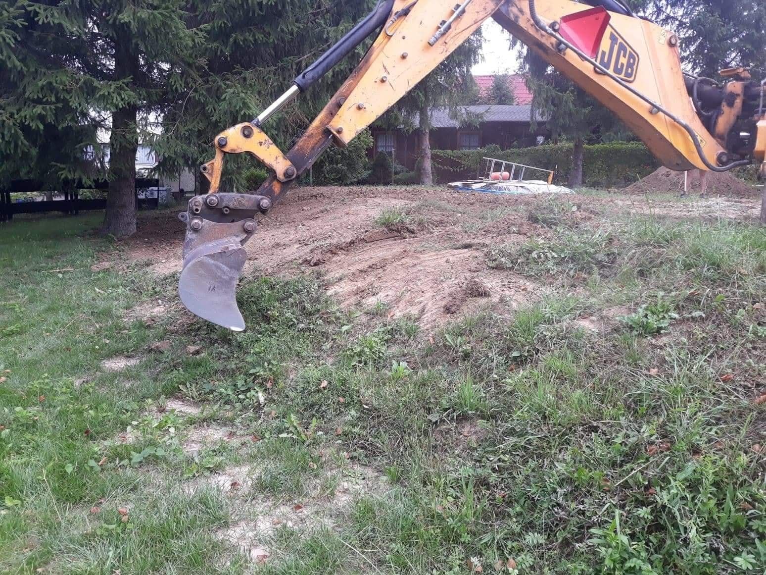 Żółta koparka JCB w trakcie prac ziemnych w Olsztynie. Widoczny wykop i fragment domu w tle. Prace ziemne z bliska, zieleń i drzewa w otoczeniu.