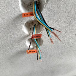 Marek Zych - Przygotowane kable zasilające, wystające z trzech otworów w ścianie, oznaczone pomarańczowymi etykietami z napisem 'Gniazdo', 'Zmywarka' i 'Lodówka'.