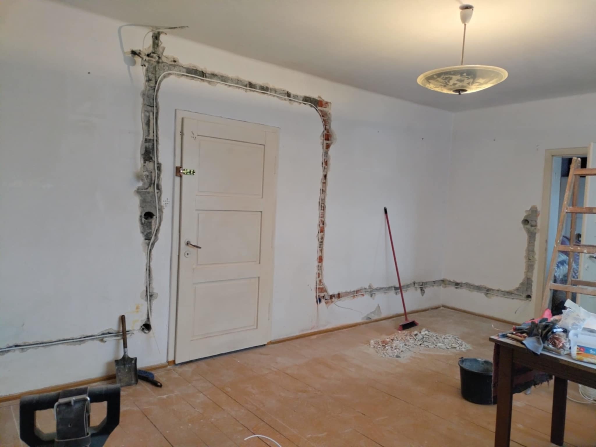 Remont instalacji elektrycznej w pokoju: kable w bruzdach na ścianach wokół drzwi, narzędzia, resztki gruzu na podłodze. Widok na wnętrze w trakcie prac.