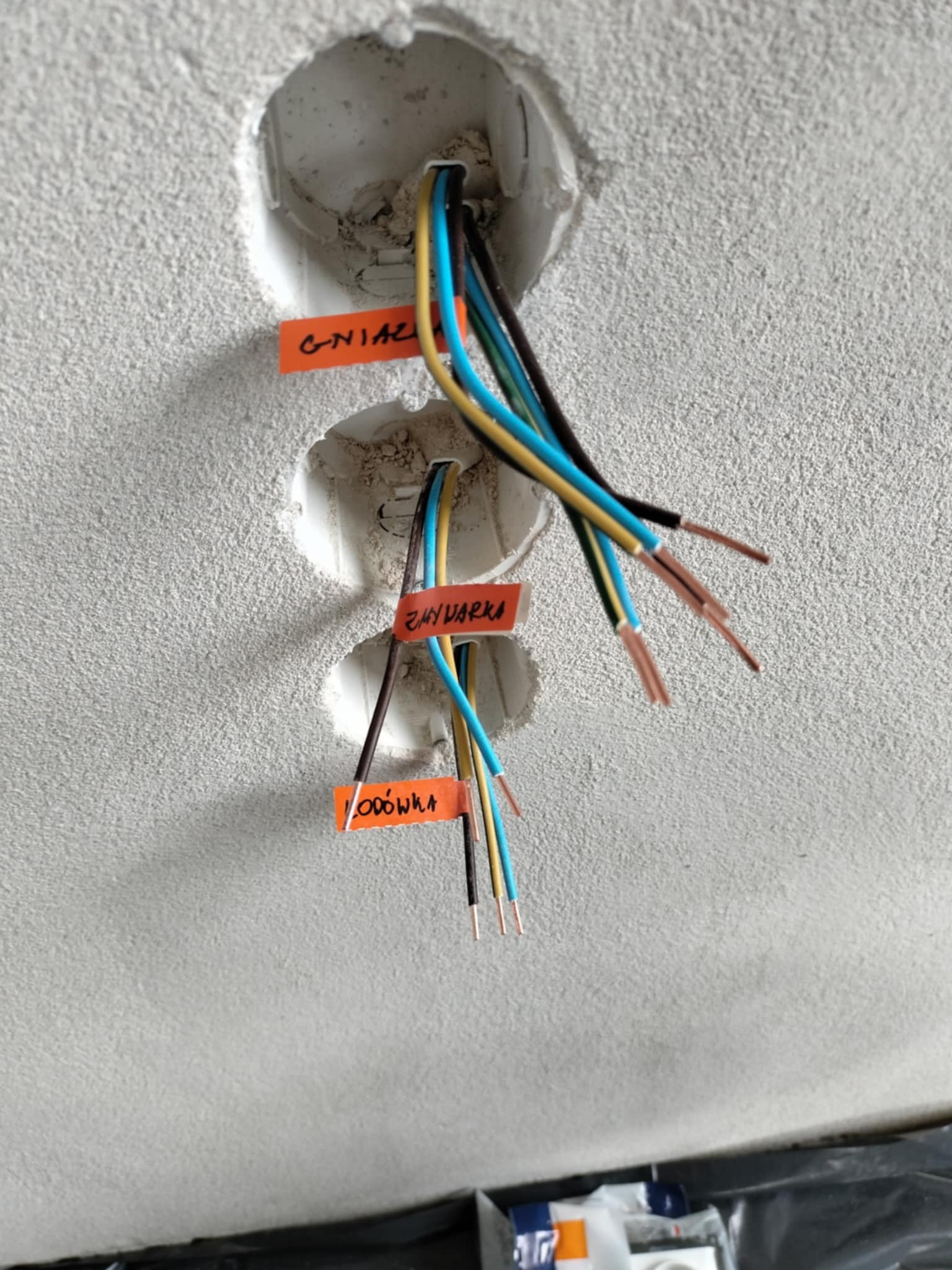 Przygotowane kable zasilające, wystające z trzech otworów w ścianie, oznaczone pomarańczowymi etykietami z napisem 'Gniazdo', 'Zmywarka' i 'Lodówka'.