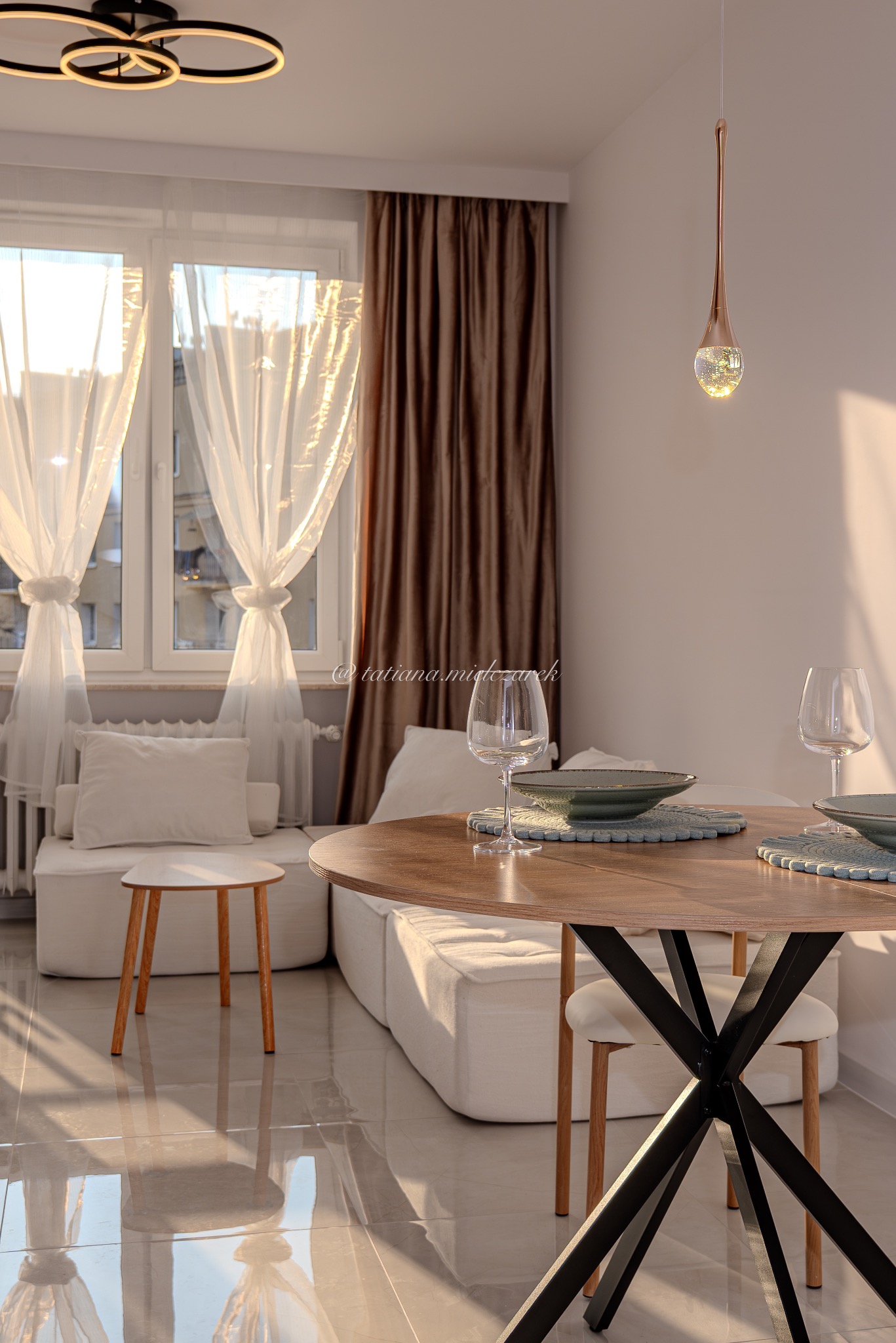 Jasne wnętrze z okrągłym stołem, nakrytym do posiłku. W tle biała sofa modułowa i okno z delikatnymi zasłonami. Ciepłe światło słoneczne podkreśla detale.