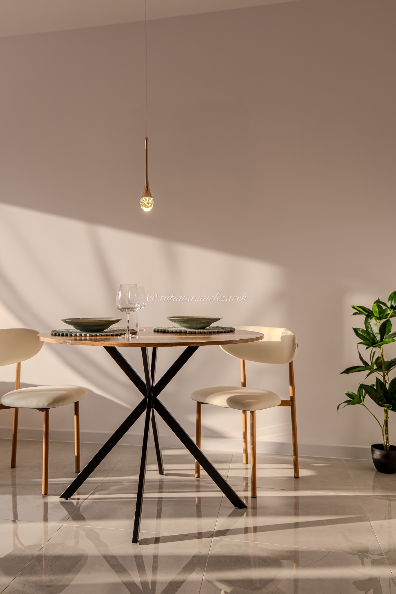 Minimalistyczna aranżacja jadalni: okrągły stół z czarnymi nogami, dwa krzesła, zastawa i roślina doniczkowa w ciepłym świetle słonecznym.