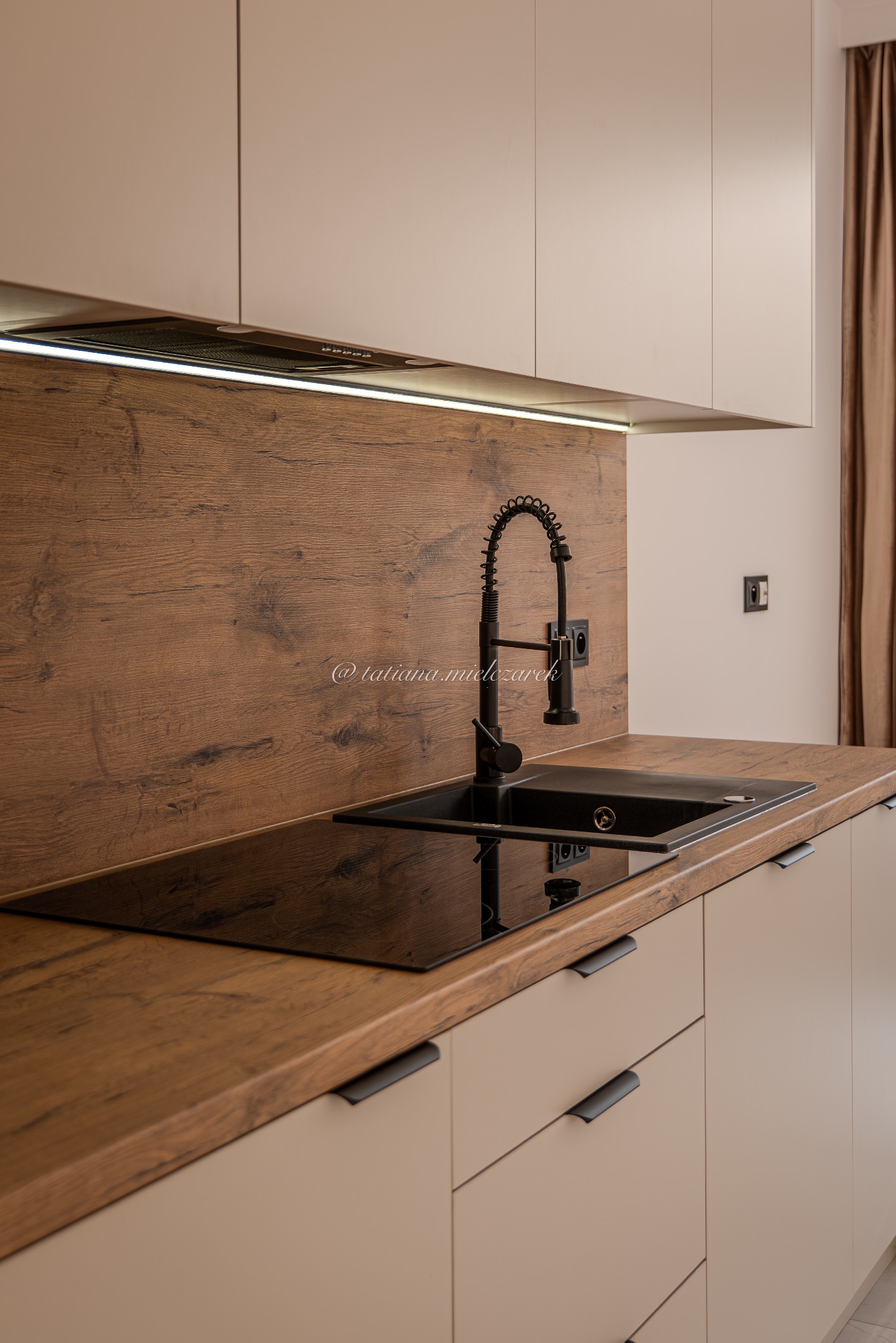 Nowoczesna kuchnia z drewnianym blatem i czarną armaturą. Minimalistyczne szafki w beżowym odcieniu. Ujęcie na zlew i płytę indukcyjną.