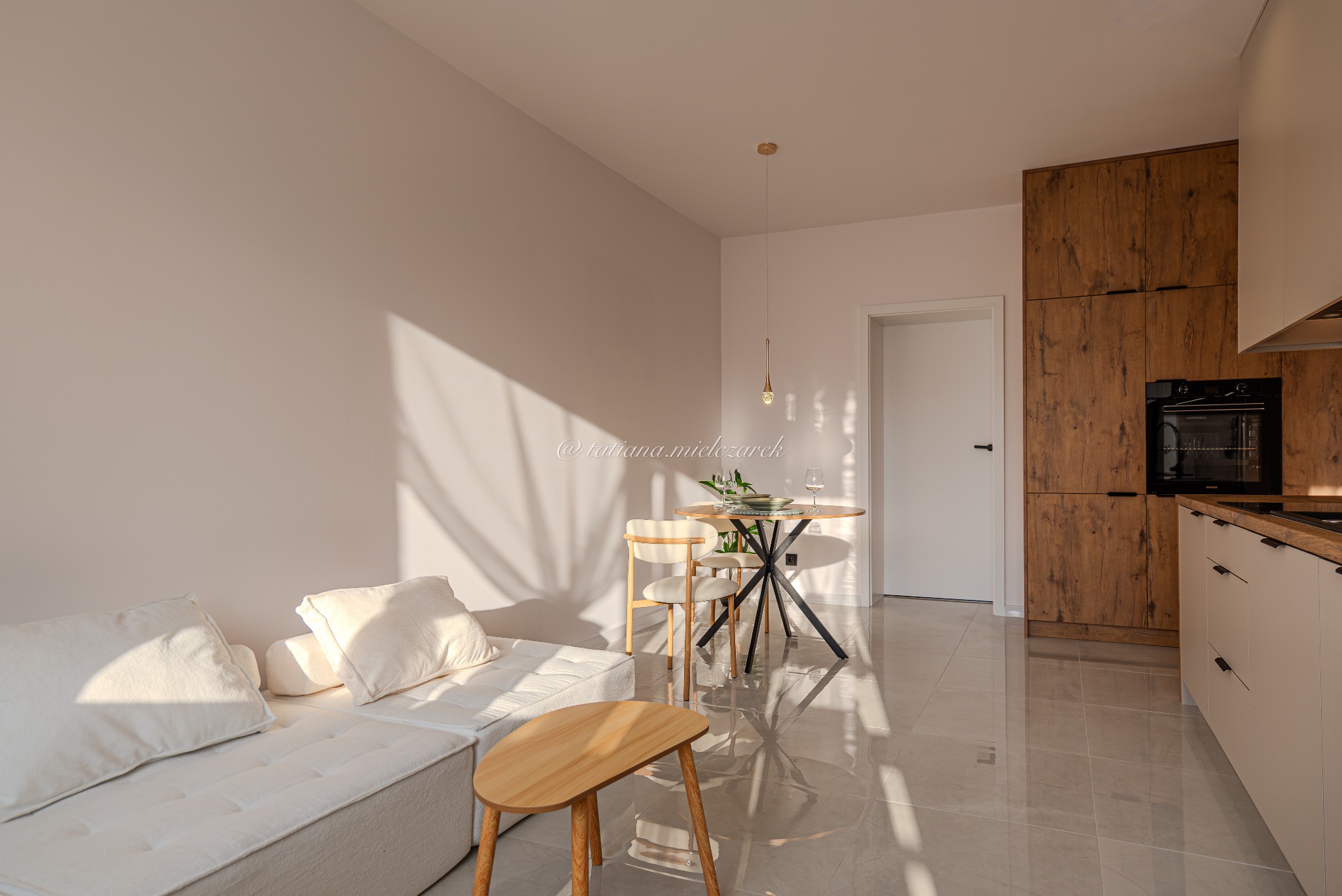 Jasne wnętrze: sofa, stół z krzesłami i zabudowa kuchenna z drewnianymi frontami. Na podłodze połyskujące płytki. Promienie słońca wpadają przez okno.