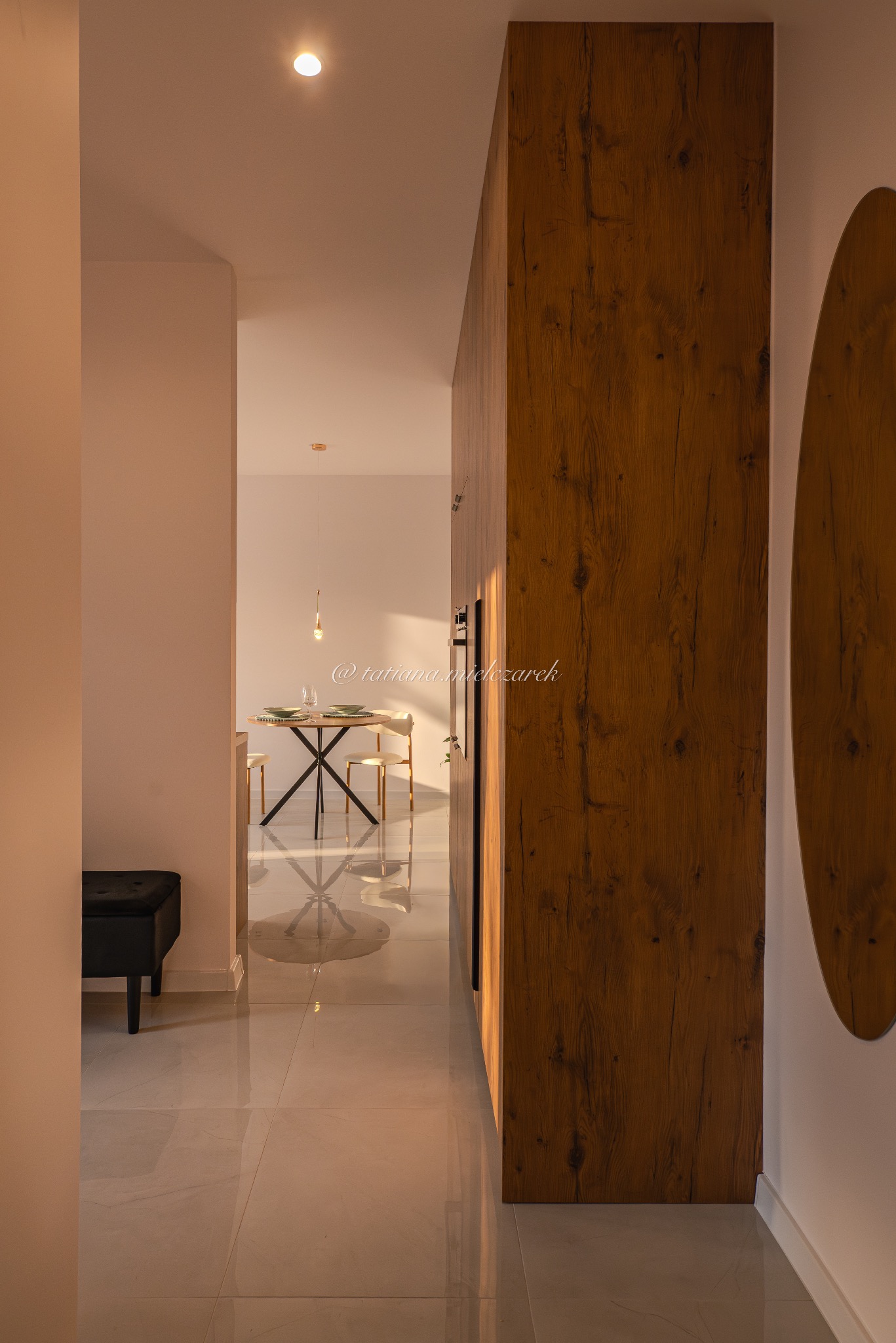 Wnętrze mieszkania z perspektywy korytarza: widok na jadalnię z okrągłym stołem i drewnianą zabudową. Ciepłe światło podkreśla fakturę drewna i połysk płytek.