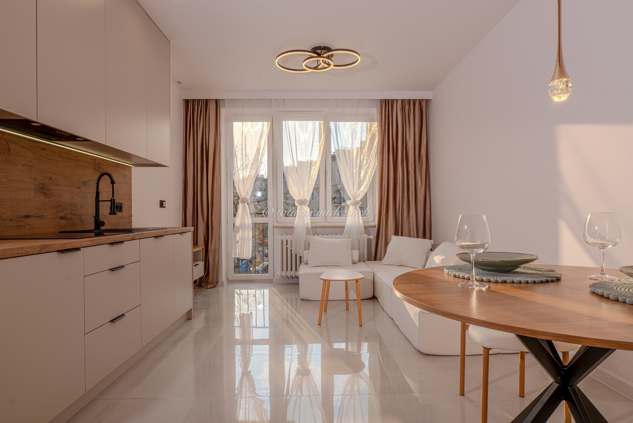 Jasne wnętrze z aneksem kuchennym i okrągłym stołem w Częstochowie. Minimalistyczny design, beżowe szafki, drewniany blat i parkiet. Widok na okno z firanami.