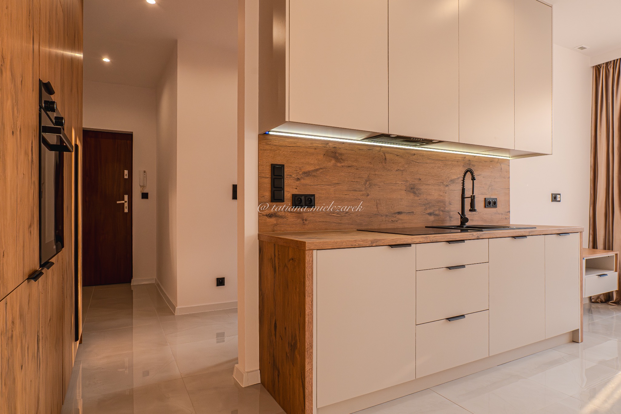 Nowoczesna kuchnia z beżowymi szafkami i drewnianym blatem. Czarna bateria i płyta indukcyjna. Widoczny fragment przedpokoju z drewnianą zabudową i drzwiami.