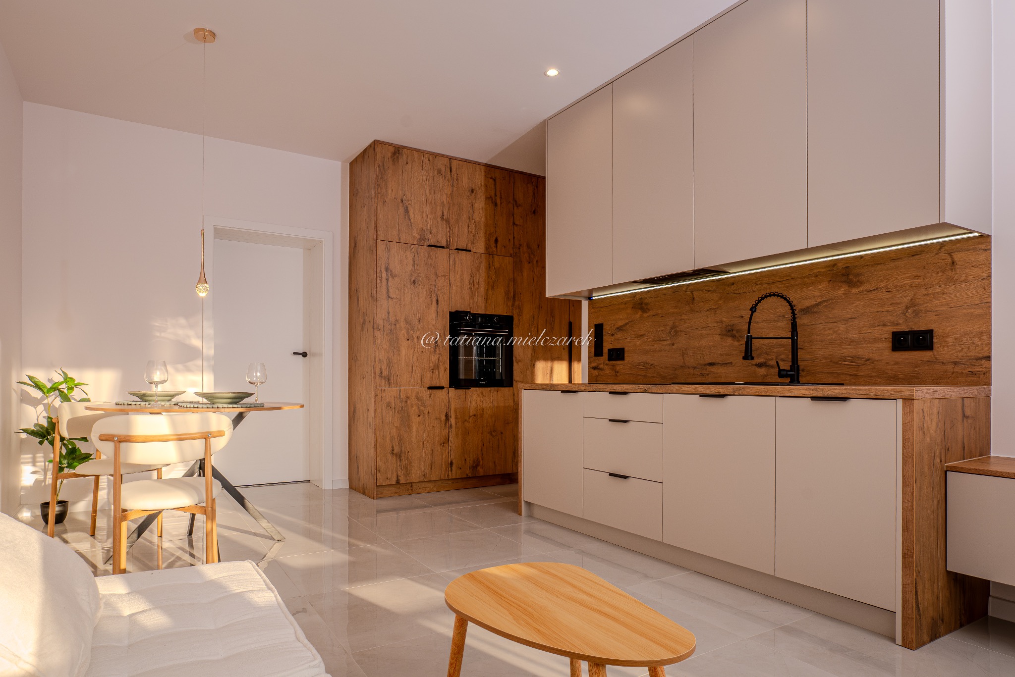 Jasne wnętrze z drewnianą zabudową kuchenną i beżowymi szafkami. Stół z nakryciem i krzesłami, sofa. Podłoga z połyskiem. Widoczne oświetlenie podszafkowe.