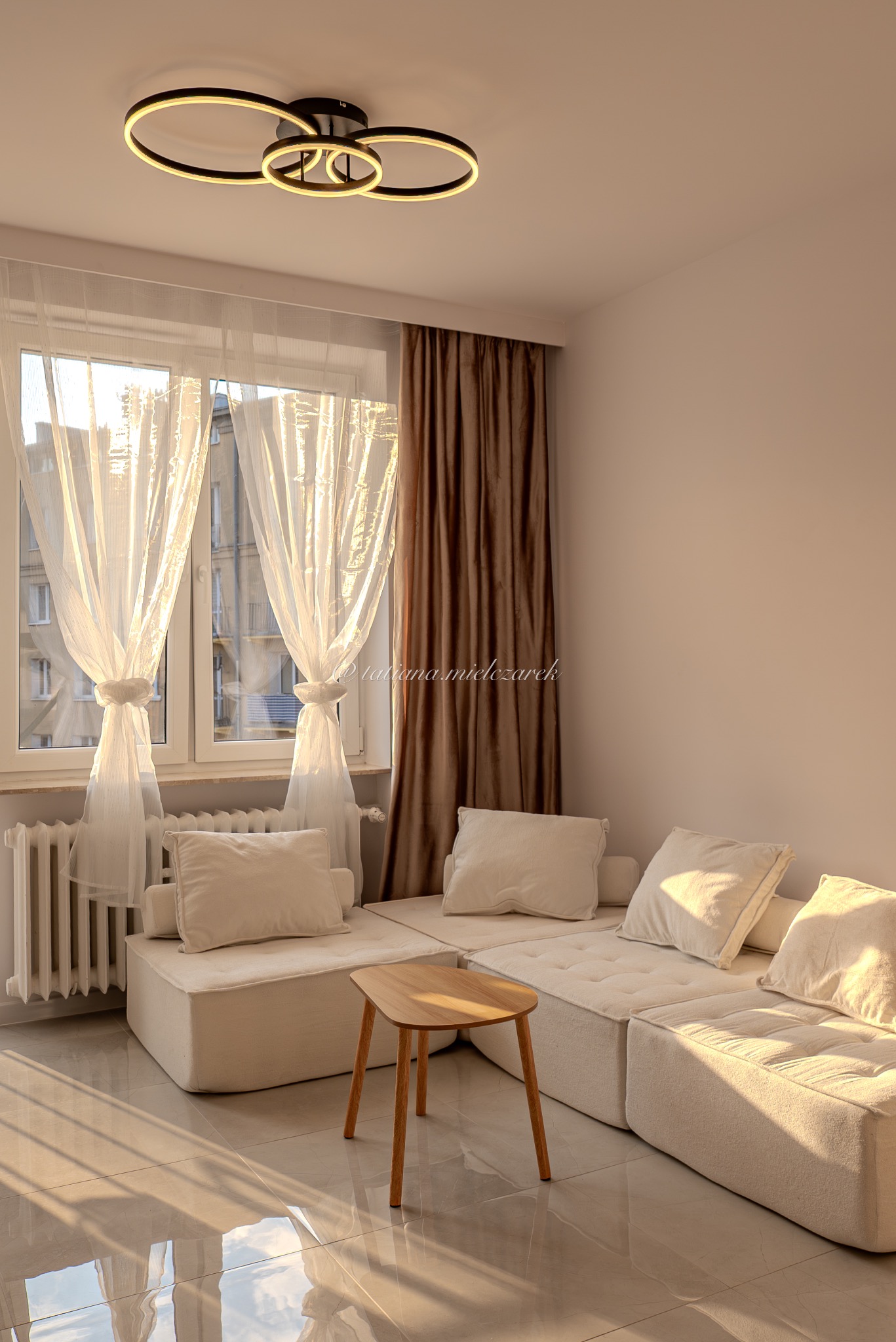 Jasne wnętrze z modułową sofą, drewnianym stolikiem i nowoczesną lampą sufitową. Okno z białymi firanami wpuszcza ciepłe światło słoneczne, odbijające się w polerowanej podłodze.