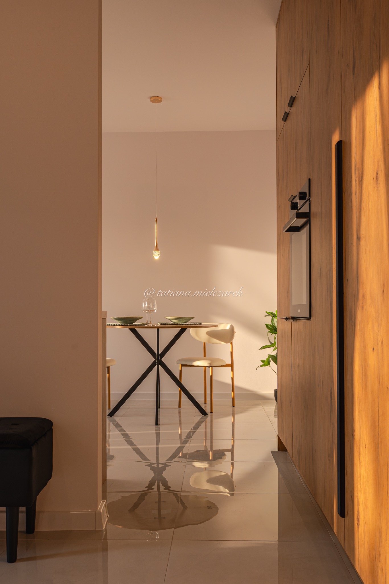 Nowoczesna jadalnia z okrągłym stołem na metalowej podstawie, krzesłami i wiszącą lampą. Widok z korytarza, błyszcząca podłoga odbija światło. Drewniana zabudowa kuchenna po prawej.