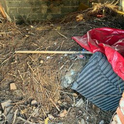 CASZAP - Zrujnowane wnętrze z gruzem, gałęziami, starym materacem i czerwoną folią. Widoczne ceglane ściany i okno w tle. Zaniedbane pomieszczenie przed sprzątaniem.