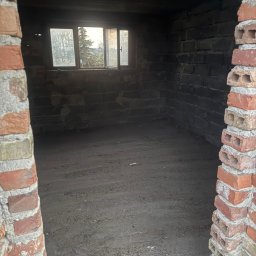 CASZAP - Ciemne, ceglane wnętrze piwnicy z oknem i ziemią na podłodze, widziane z zewnątrz przez ceglane obramowanie. Widoczne ślady wilgoci i zabrudzeń.