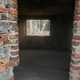 CASZAP - Surowe wnętrze w trakcie remontu: ceglane filary, okno z widokiem na brzozy, betonowa podłoga. Widok na zewnątrz przez okno w surowej ścianie.