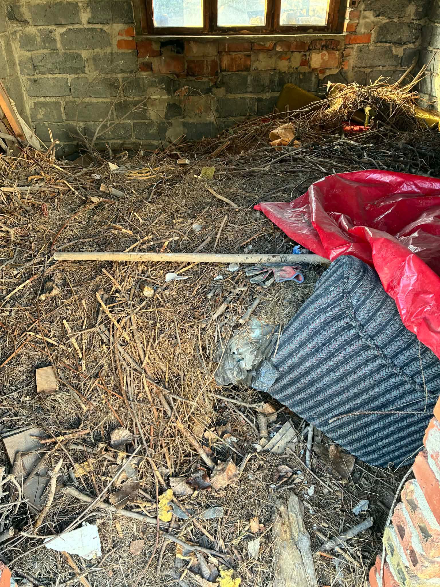 Zrujnowane wnętrze z gruzem, gałęziami, starym materacem i czerwoną folią. Widoczne ceglane ściany i okno w tle. Zaniedbane pomieszczenie przed sprzątaniem.