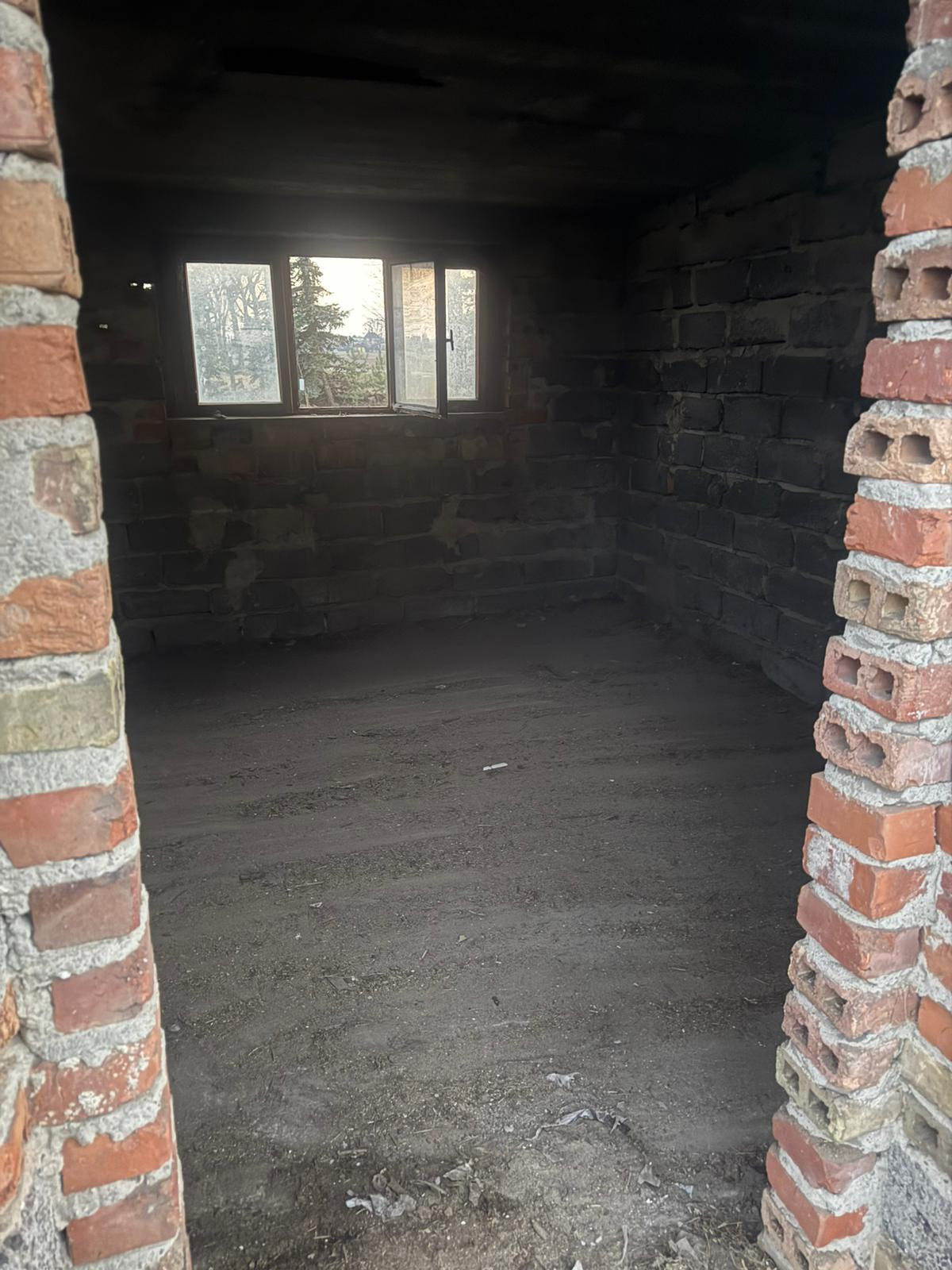 Ciemne, ceglane wnętrze piwnicy z oknem i ziemią na podłodze, widziane z zewnątrz przez ceglane obramowanie. Widoczne ślady wilgoci i zabrudzeń.