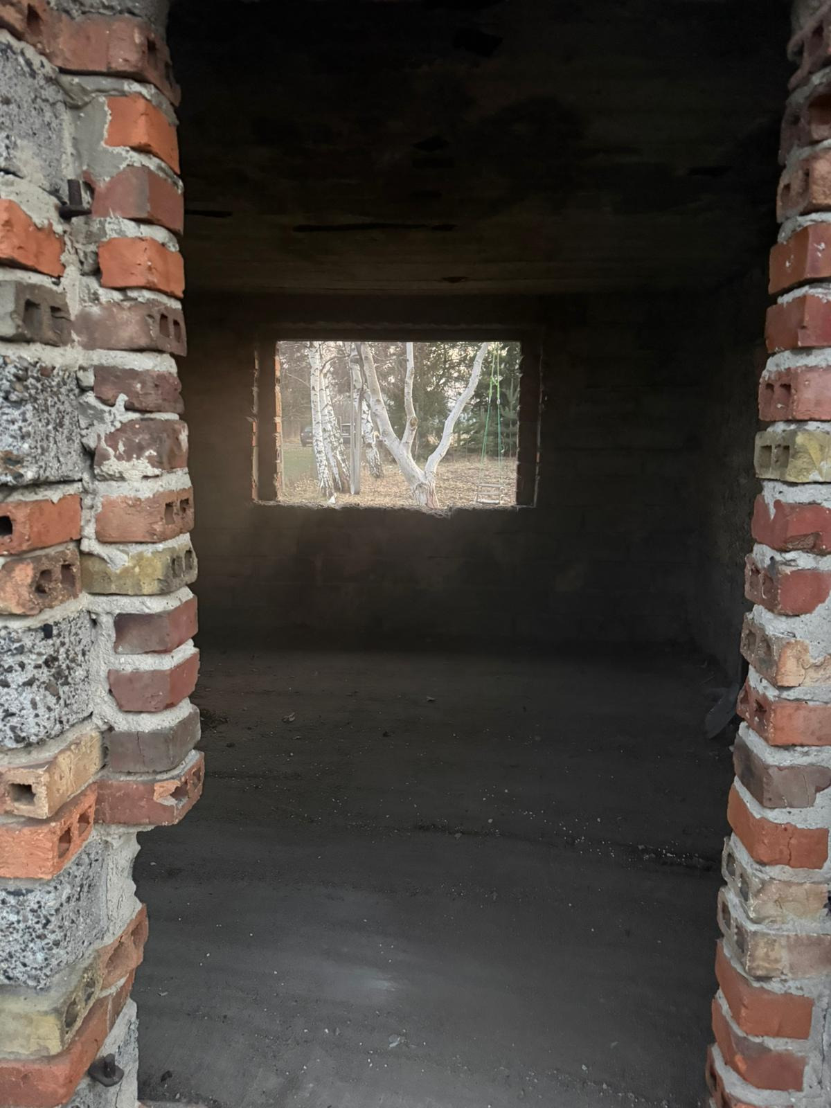 Surowe wnętrze w trakcie remontu: ceglane filary, okno z widokiem na brzozy, betonowa podłoga. Widok na zewnątrz przez okno w surowej ścianie.