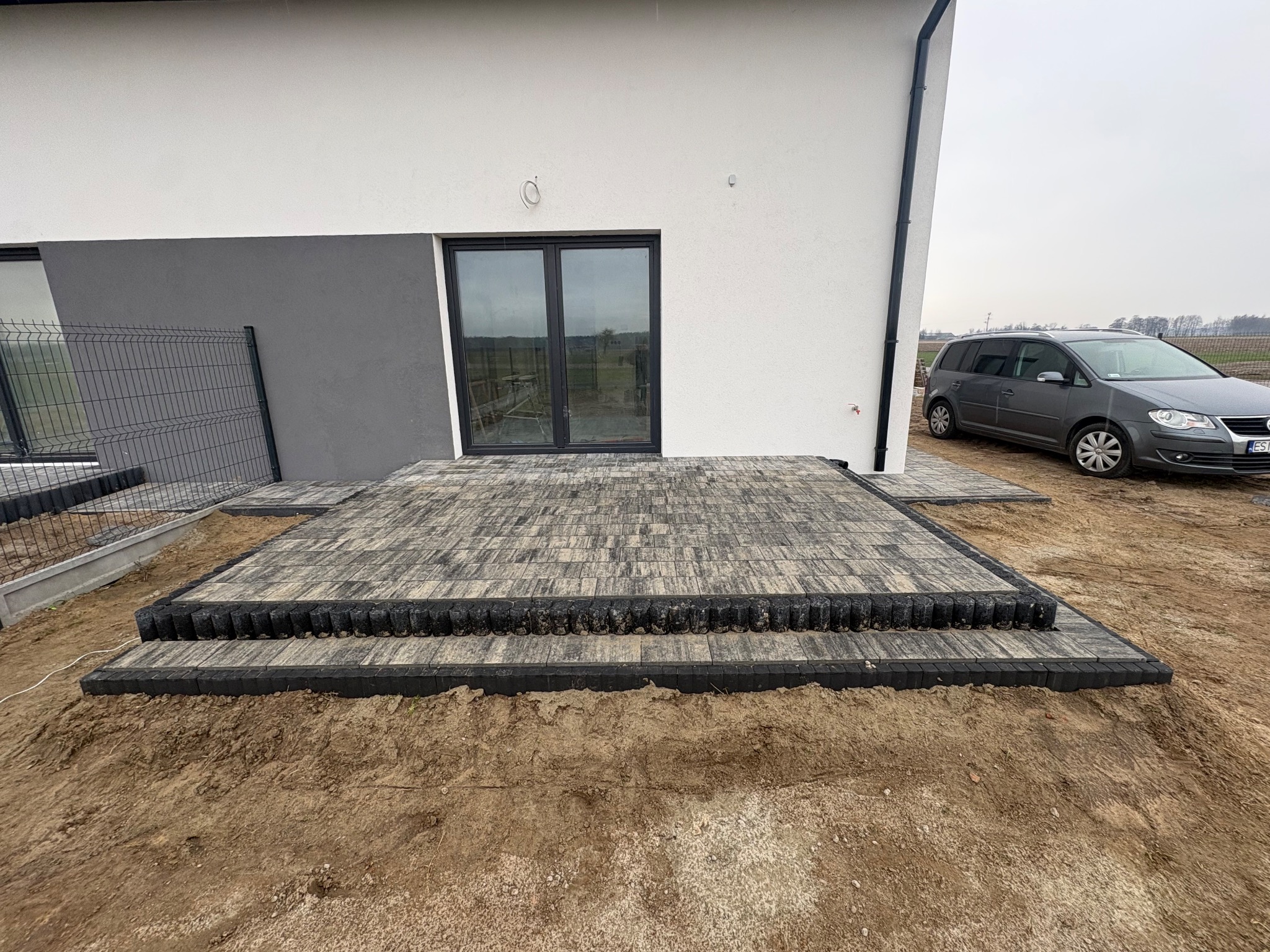 Ułożona kostka brukowa tworząca taras przy domu z szarą elewacją. Widoczny fragment ogrodzenia i samochód. Kostka w odcieniach szarości i czerni.