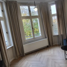 Ozdoba okna - Beżowe zasłony w eleganckim salonie z parkietem w jodełkę i dużymi oknami. Widok na zieleń za oknem. Minimalistyczna lampa sufitowa.