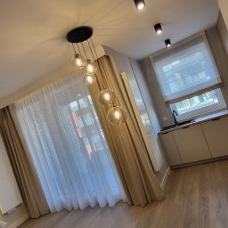 Ozdoba okna - Jasne wnętrze z roletami rzymskimi w oknie, zwiewnymi zasłonami i nowoczesnym oświetleniem sufitowym. Parkiet na podłodze.