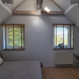 Ozdoba okna - Jasna sypialnia na poddaszu z dwoma oknami przesłoniętymi drewnianymi żaluzjami. Widoczny fragment łóżka z szarym zagłówkiem i drewniana belka stropowa.