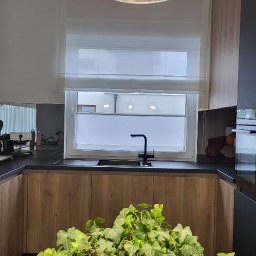 Ozdoba okna - Jasna roleta rzymska zamontowana w oknie nowoczesnej kuchni z drewnianymi szafkami i czarnym blatem. Na pierwszym planie roślina doniczkowa.