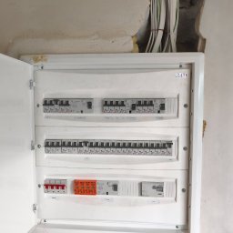 Piotr Żebrowski - Otwarta biała rozdzielnica elektryczna z rzędami szarych wyłączników nadprądowych i pomarańczowymi ogranicznikami przepięć, zamontowana w nieotynkowanej ścianie z widocznymi przewodami.