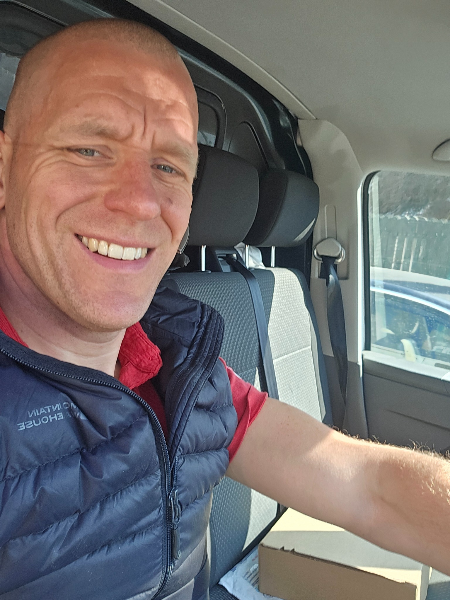 Uśmiechnięty mężczyzna w kamizelce siedzi w samochodzie dostawczym, obok niego karton. Widok z perspektywy kierowcy, jasne światło dzienne.