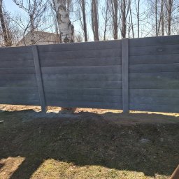 Usługi Ogulno Budowlane - Nowoczesne, szare ogrodzenie panelowe z betonu imitującego drewno, z widocznymi słupkami, na tle drzew i budynku w oddali, w słoneczny dzień.