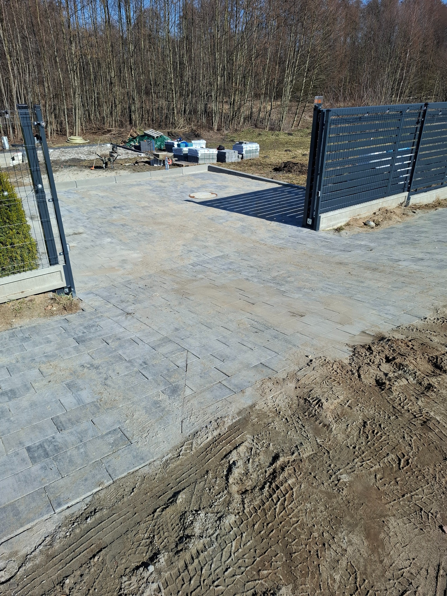 Ułożona kostka brukowa w szarym odcieniu, z widoczną bramą wjazdową i fragmentem ogrodzenia panelowego na tle lasu. Częściowo widoczne ślady opon na ziemi.