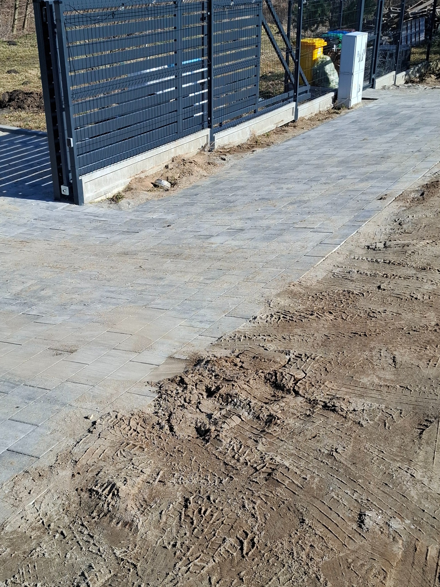 Ułożona szara kostka brukowa przy metalowym ogrodzeniu panelowym. Widoczne ślady opon na ziemi obok kostki. Częściowo ukończona realizacja.