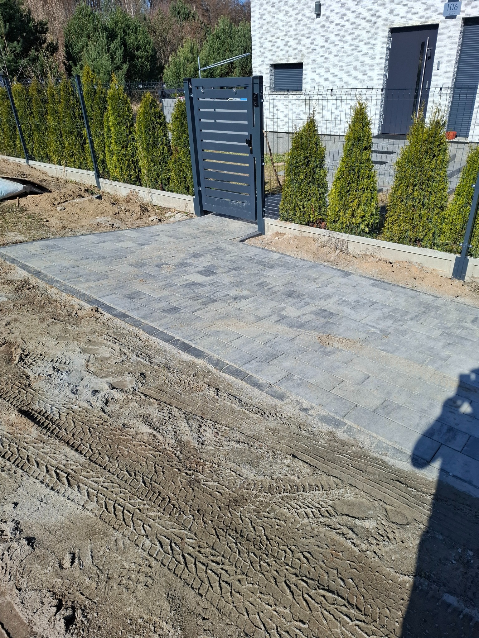 Ułożona szara kostka brukowa prowadząca do furtki z metalowym ogrodzeniem panelowym. W tle rząd tuj i budynek z numerem 106.