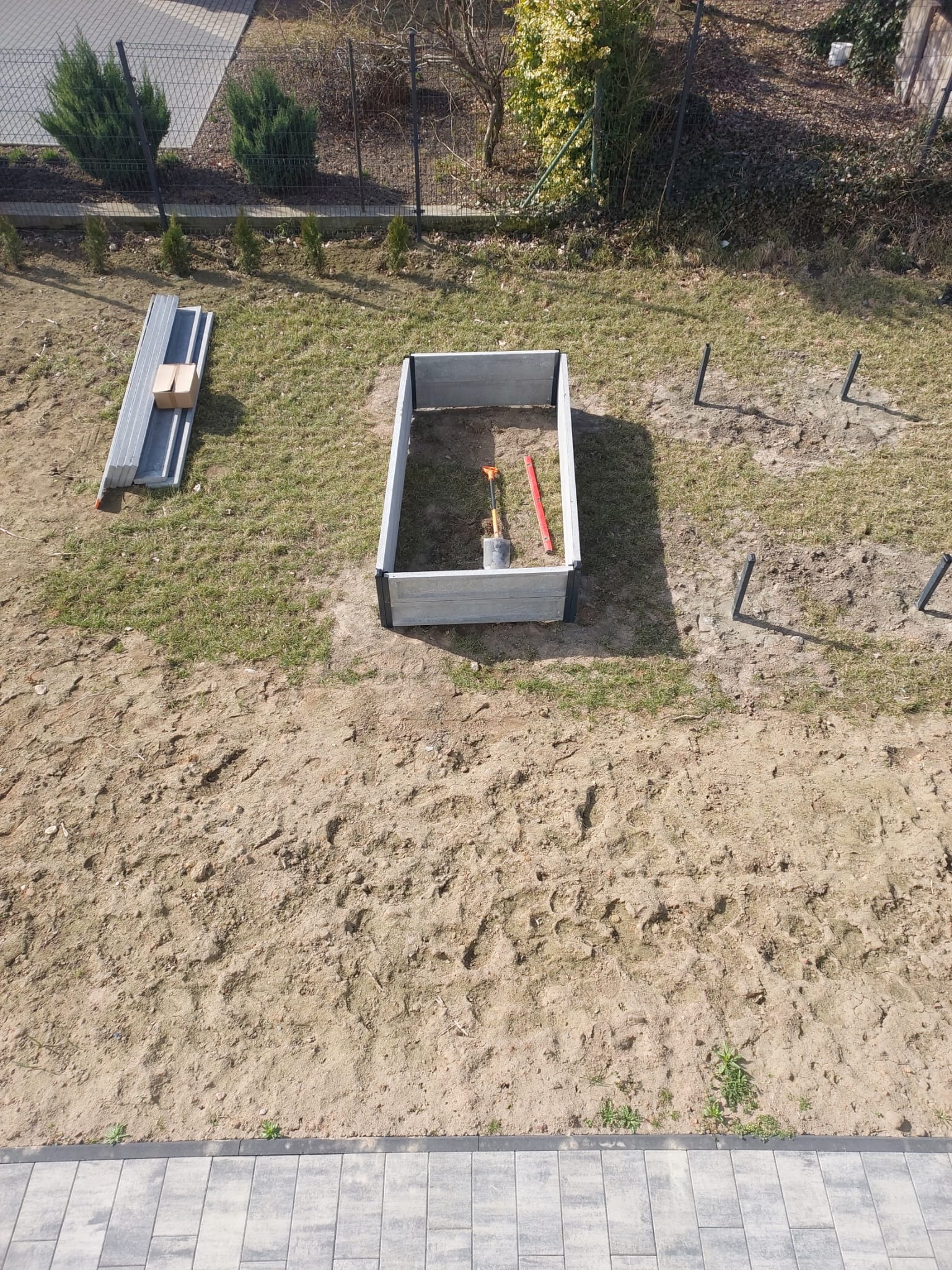 Przygotowanie podwyższonej rabaty w ogrodzie: rama z desek, narzędzia (szpadel, poziomica) w środku, obok leżą profile. Widoczne słupki ogrodzeniowe.