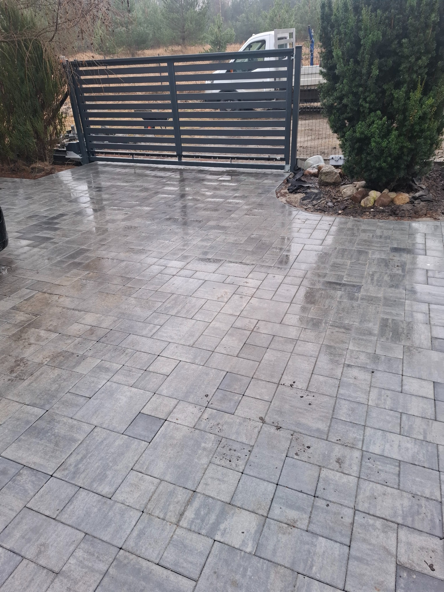 Nowoczesna, pozioma brama wjazdowa z szarych paneli, widoczna zza ułożonej kostki brukowej w odcieniach szarości. W tle zieleń drzew i częściowo widoczny biały samochód.