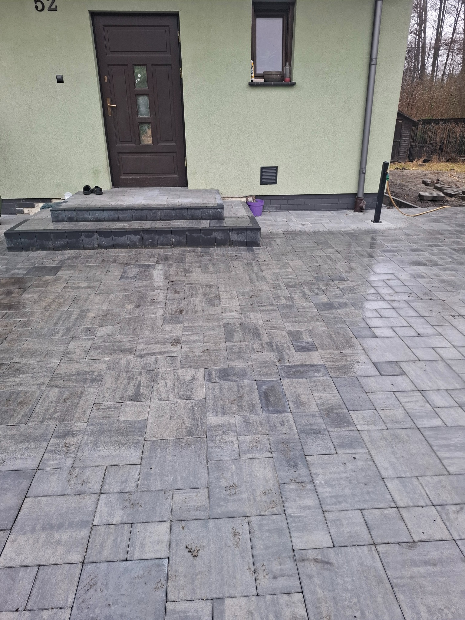 Ułożona szara kostka brukowa przed domem z trzema schodami wejściowymi. Widoczny fragment elewacji i rynna spustowa. Częściowo mokra nawierzchnia.