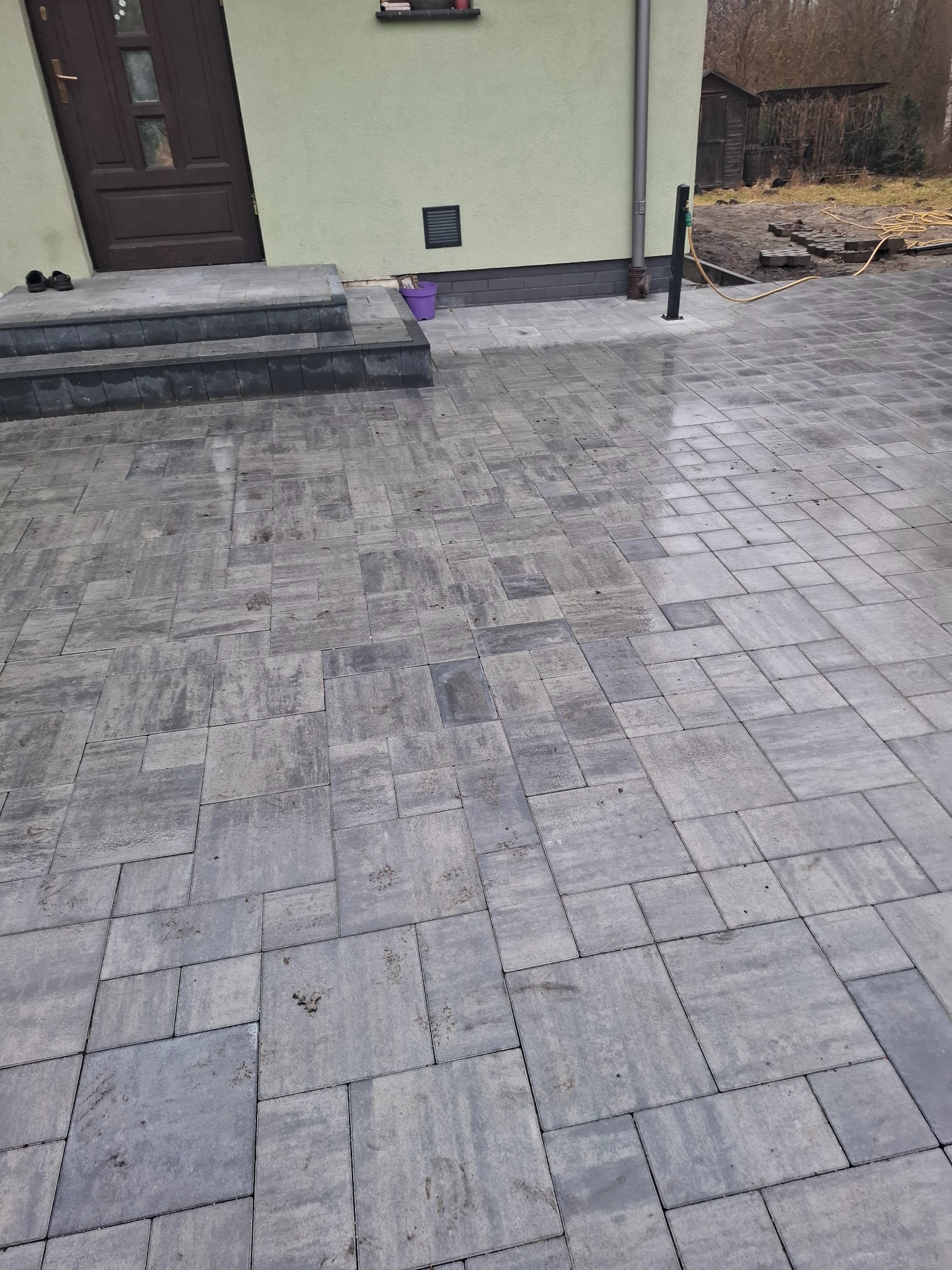 Ułożona, szara kostka brukowa przed domem z ciemnymi drzwiami i schodami. W tle widoczny fragment budynku i elementy ogrodu. Kostka o nieregularnym wzorze.