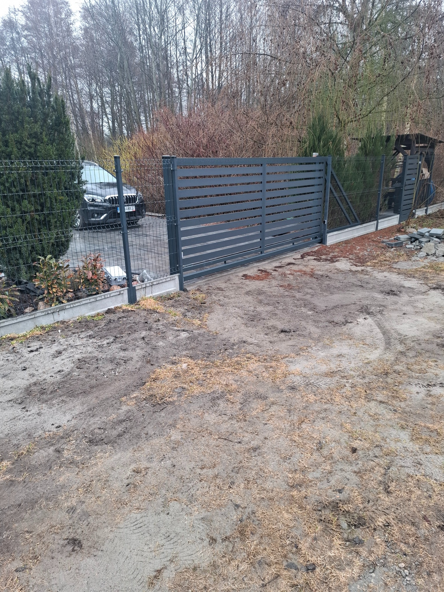 Nowoczesna, przesuwna brama w kolorze grafitowym z poziomymi panelami, zamontowana na posesji z ogrodzeniem z siatki. W tle widoczny samochód osobowy.