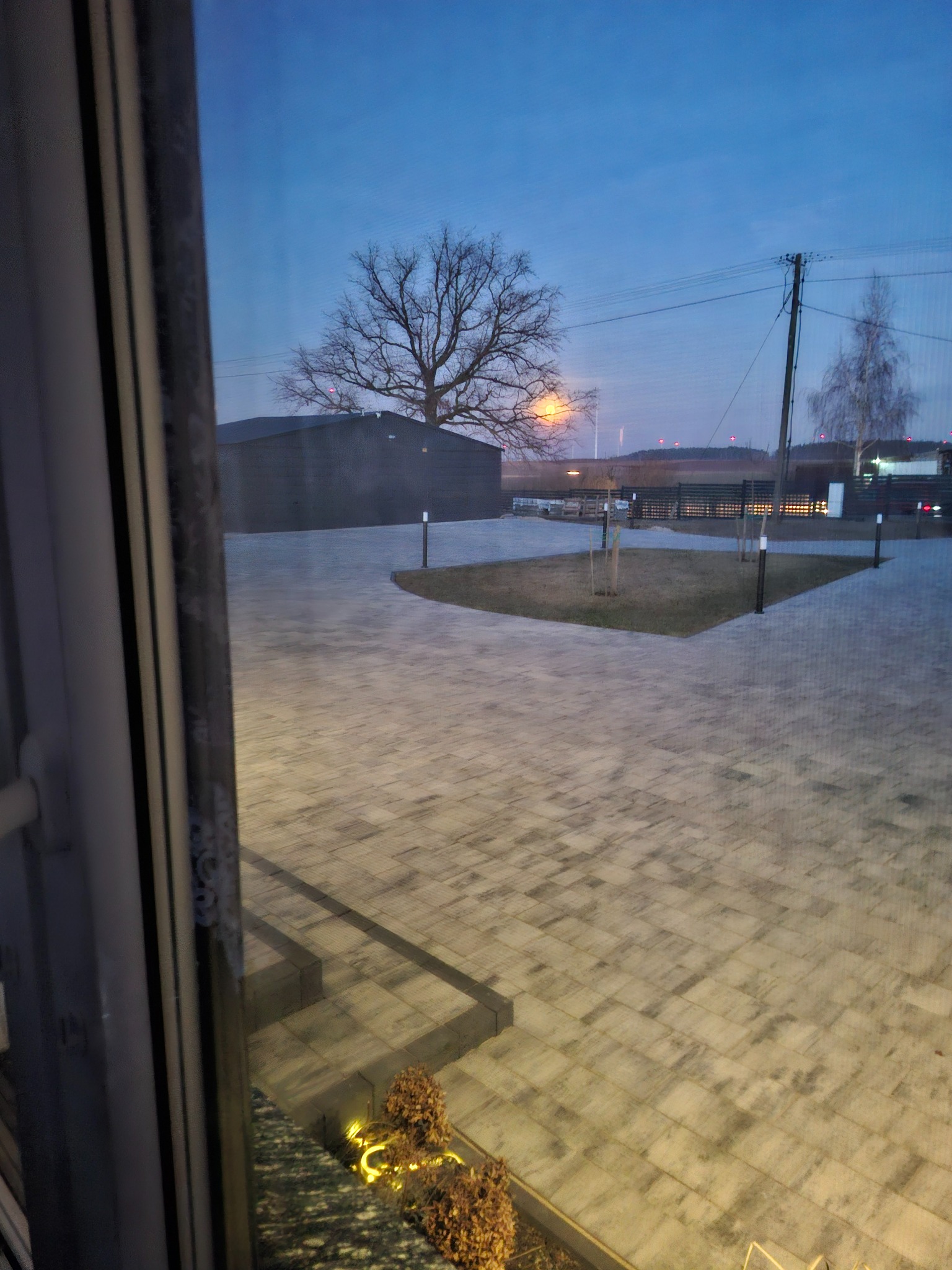 Ułożona kostka brukowa w szarym odcieniu, widok z okna na posesję z drzewami i budynkiem w tle, oświetlona lampami. Widoczne elementy ogrodu.