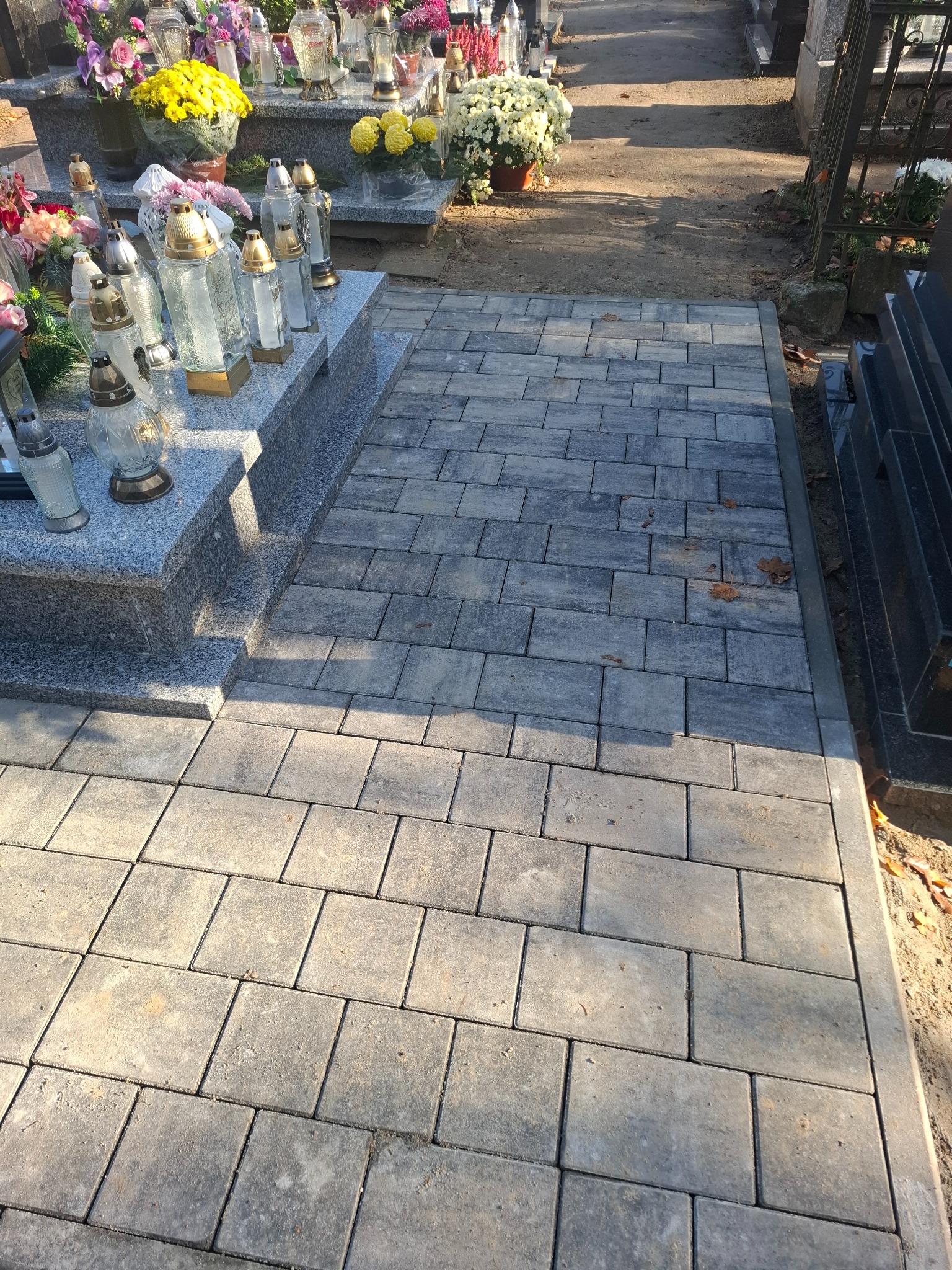 Ułożona szara kostka brukowa tworzy ścieżkę między nagrobkami na cmentarzu. Widoczne znicze i kwiaty na pomnikach. Jesienne liście na kostce.