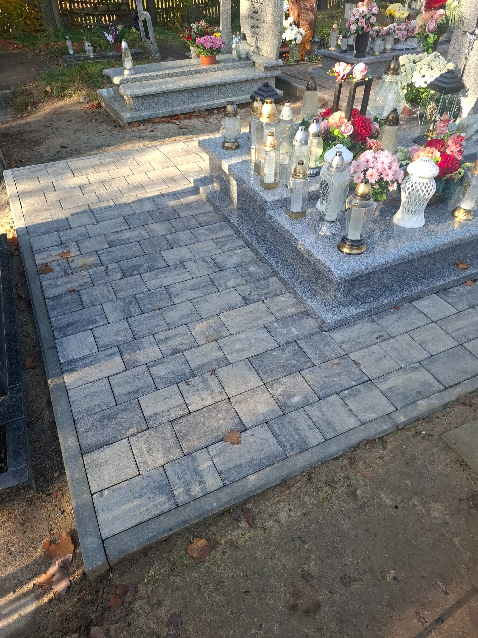Ułożona kostka brukowa w odcieniach szarości tworzy ścieżkę przy nagrobku. Widoczne znicze i kwiaty. Jesienne liście na ziemi. Całość w dziennym świetle.