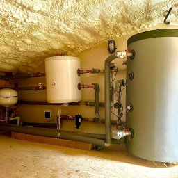 KAROLINA KAPSA - Złożona instalacja grzewcza w pomieszczeniu ze spryskaną pianą izolacyjną na suficie. Widoczne zbiorniki, rury i zawory. Całość sprawia wrażenie nowej i profesjonalnie wykonanej.