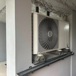 KAROLINA KAPSA - Biała pompa ciepła R32 zamontowana na szarej ścianie wewnątrz budynku. Widoczne czarne rury i izolacja piankowa na suficie. Ujęcie z dołu, lekko z lewej strony.