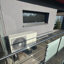 KAROLINA KAPSA - Biała pompa ciepła typu split zamontowana na zewnątrz budynku z szarą elewacją, widoczna zza metalowej balustrady. Słoneczny dzień.