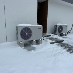 KAROLINA KAPSA - Dwie pompy ciepła Ferroli, przysypane śniegiem, stoją na betonowych podstawach przed budynkiem w zimowej scenerii. Widoczne rury i elementy instalacji.