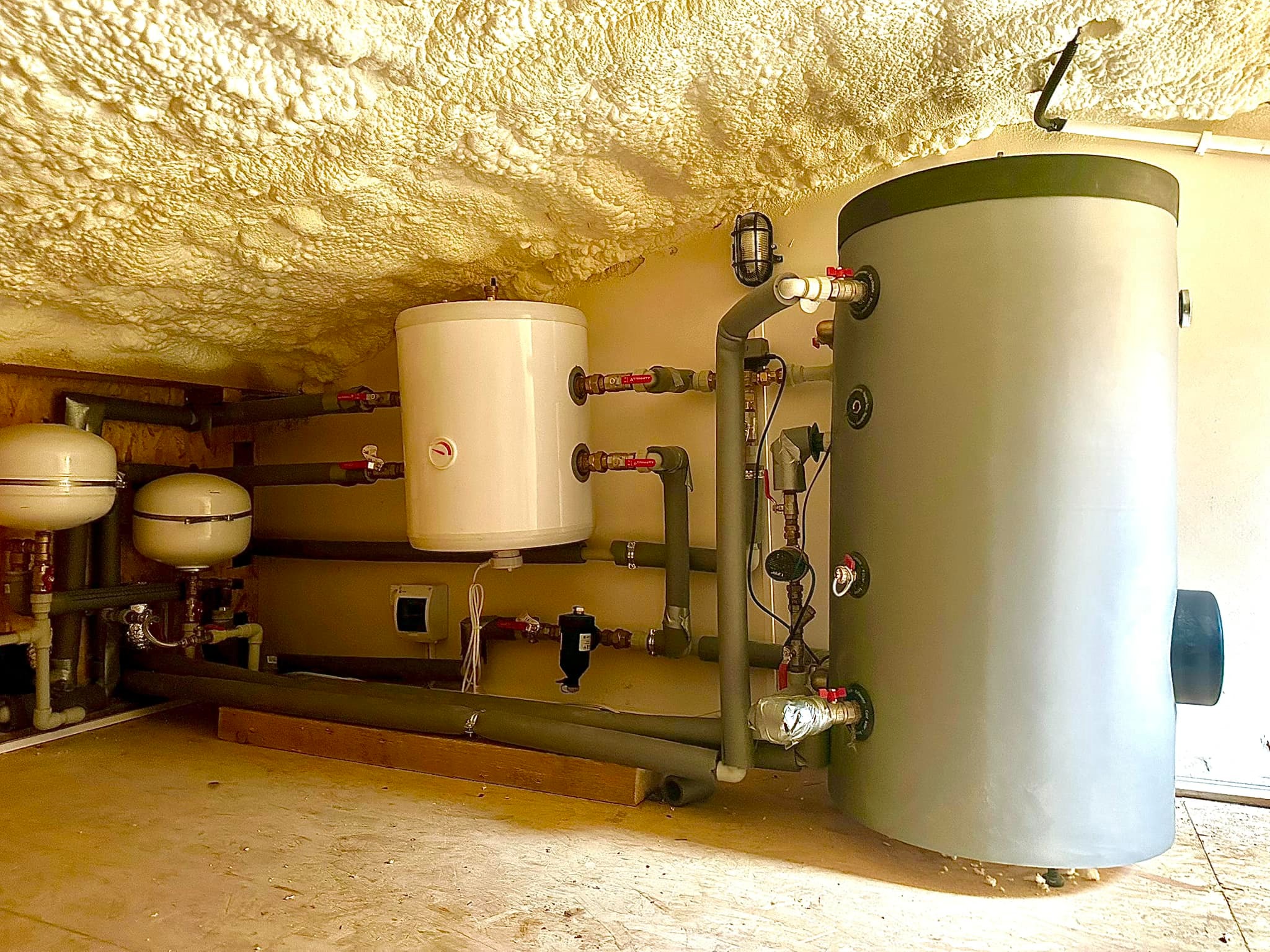 Złożona instalacja grzewcza w pomieszczeniu ze spryskaną pianą izolacyjną na suficie. Widoczne zbiorniki, rury i zawory. Całość sprawia wrażenie nowej i profesjonalnie wykonanej.