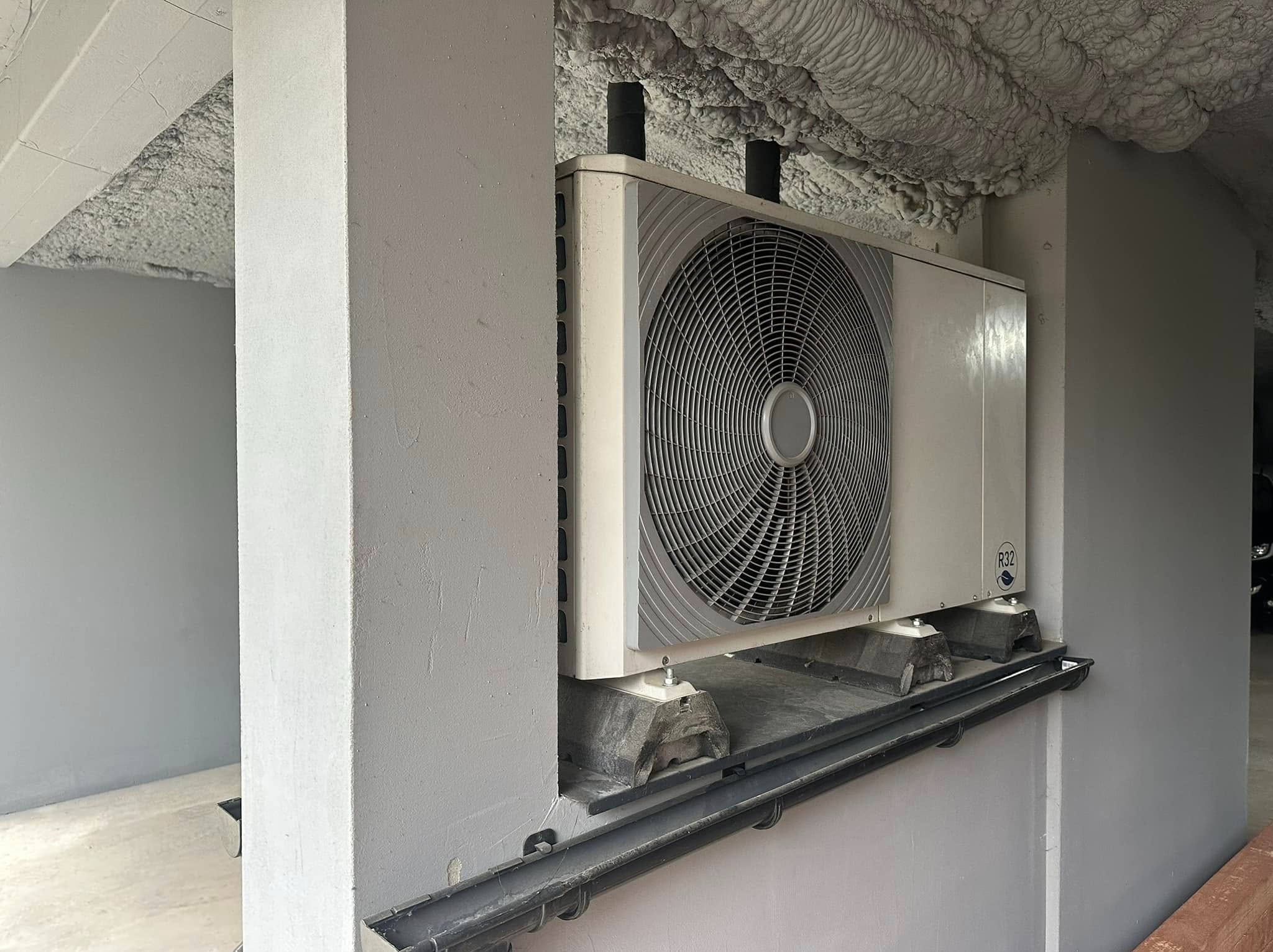 Biała pompa ciepła R32 zamontowana na szarej ścianie wewnątrz budynku. Widoczne czarne rury i izolacja piankowa na suficie. Ujęcie z dołu, lekko z lewej strony.