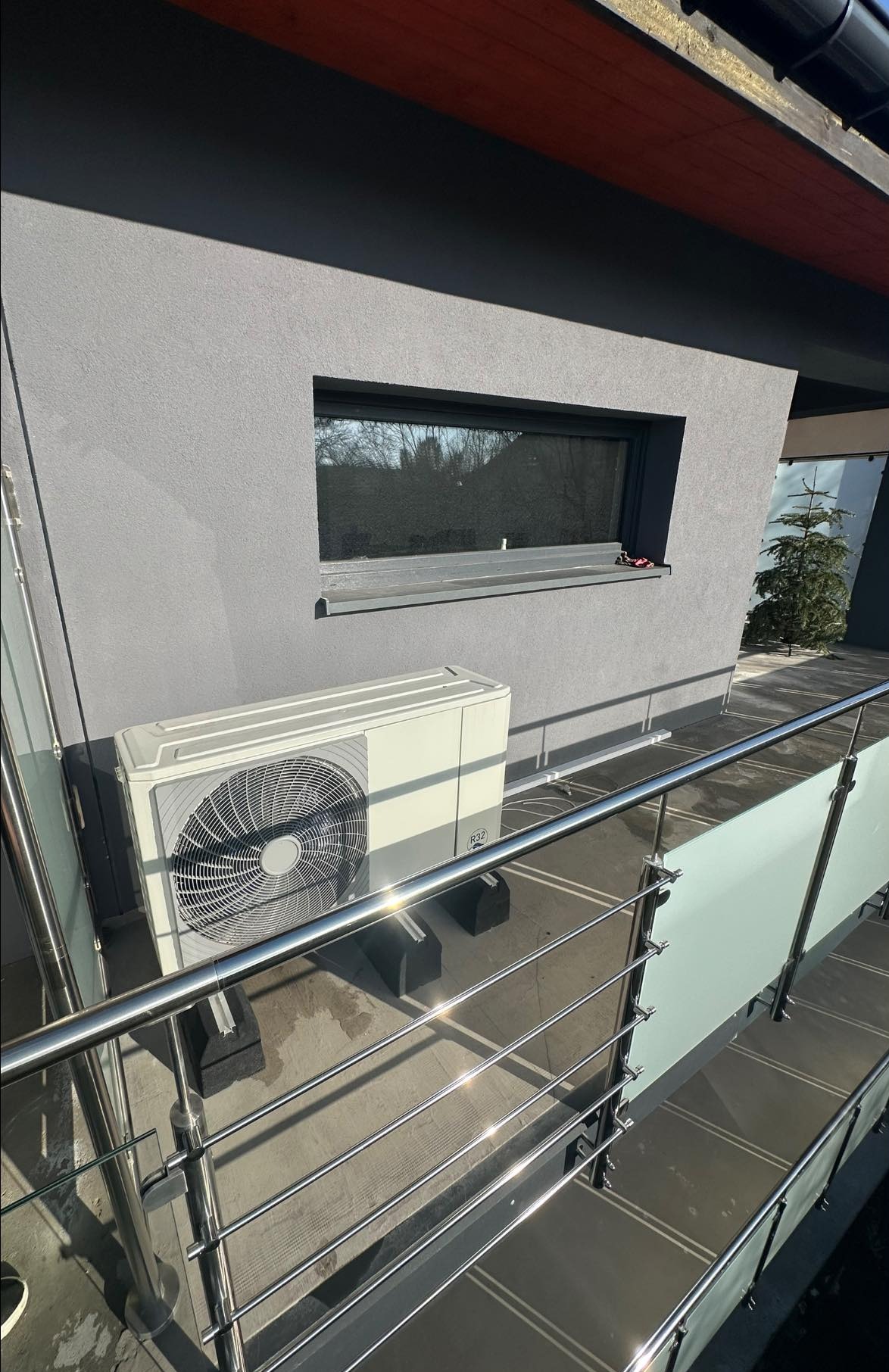 Biała pompa ciepła typu split zamontowana na zewnątrz budynku z szarą elewacją, widoczna zza metalowej balustrady. Słoneczny dzień.