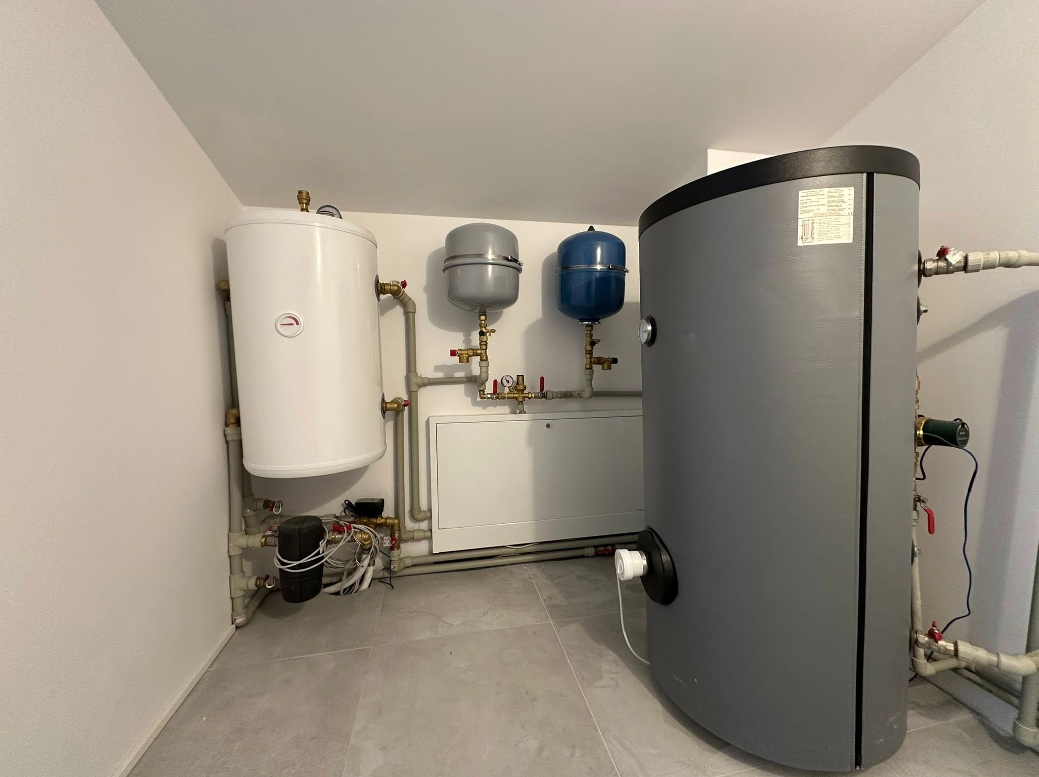 Zestaw zbiorników i instalacji w kotłowni: biały bojler, szary i niebieski zbiornik wyrównawczy, rury z zaworami. Nowoczesna instalacja w jasnym pomieszczeniu.